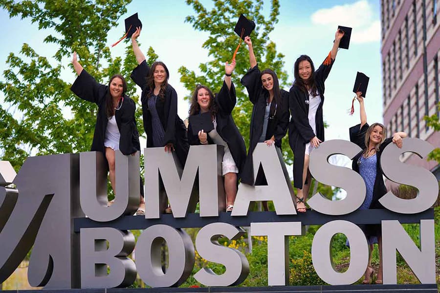 Students in graduation gowns stand and cheer at the UMass Boston sign.