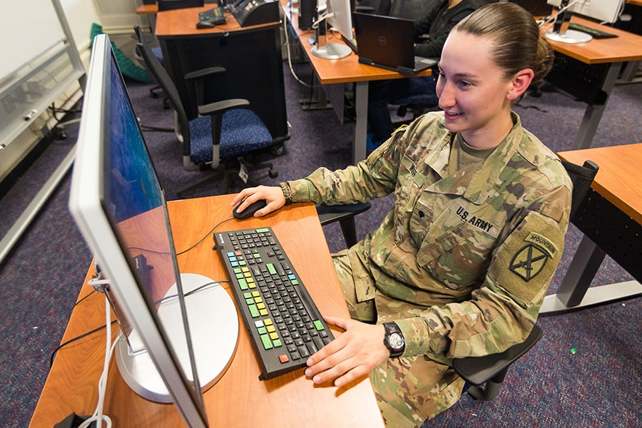 Student in fatigues smiles while working on computer inside.