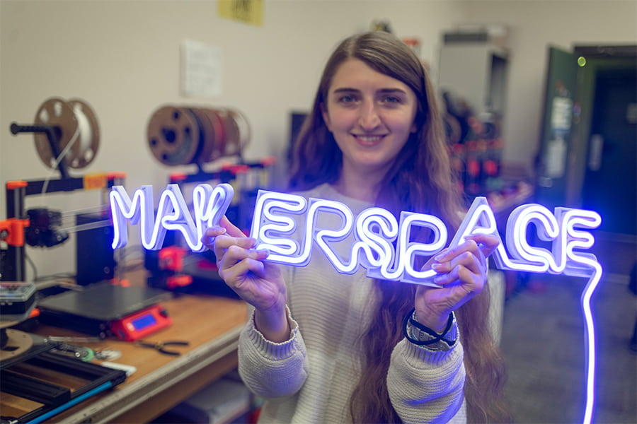 Sydney holds up 3d illuminated Makerspace sign in front of 3d printers.