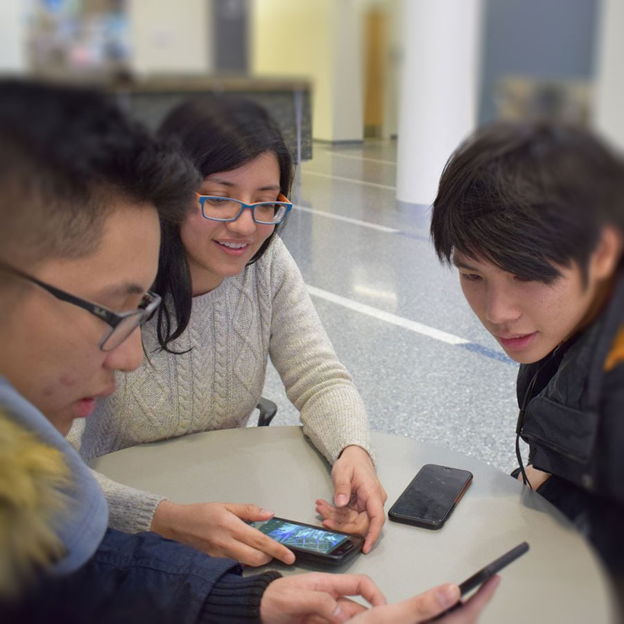 Three students sit at table in ISC looking at phone