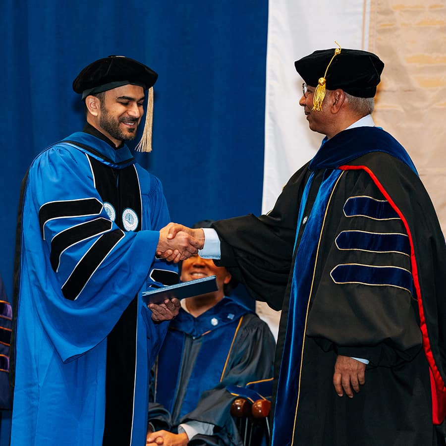 PHd student shakes hand while receiving degree at doctoral hooding ceremony.