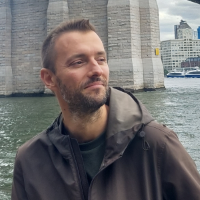 Man with short beard and short hair wearing brown jacket, standing in front of water